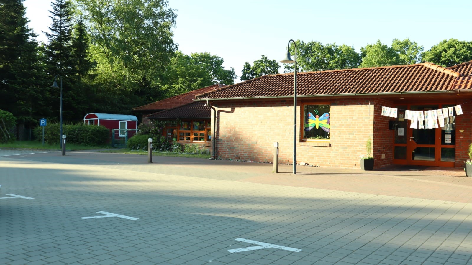 Gepflasterter Parkplatz vor einem eingeschossigen Backsteingebäude mit roten Dachziegeln, einem bunten Schmetterlingsbild im Fenster, einer Girlande über dem Eingang, Laternenpfosten und Bäumen sowie einem roten Anhänger hinter Hecken links.