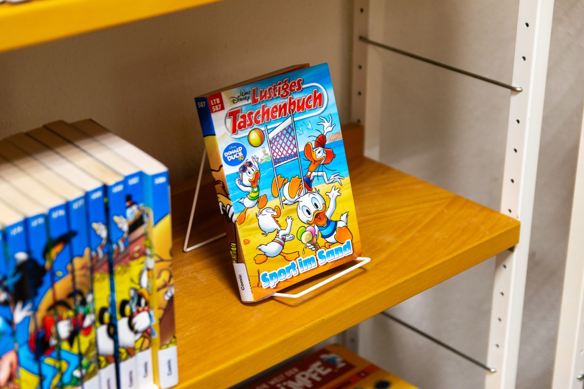 Auf einem Holzregal steht in einem weißen Drahtaufsteller ein farbenfrohes „Lustiges Taschenbuch“ mit Donald-Duck-Figuren, die am Strand Volleyball spielen; links sind weitere Comicbände sichtbar.