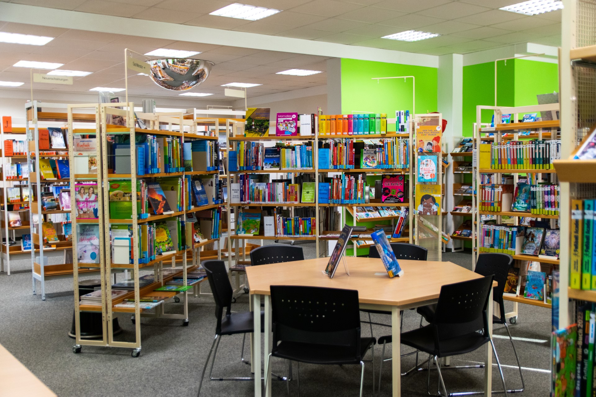 Innenraum einer Bibliothek mit mehreren Reihen offener Bücherregale voller farbiger Bücher vor einer grünen Wand; im Vordergrund ein sechseckiger Holztisch mit vier schwarzen Stühlen und aufgestellten Büchern.