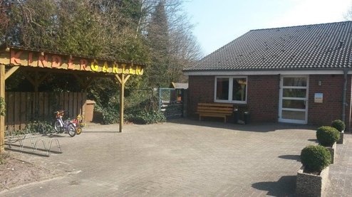 Gepflasterter Vorplatz eines eingeschossigen Backsteingebäudes mit Eingangstür und Sitzbank; links ein überdachter Holzunterstand mit bunten Buchstaben und zwei Kinderfahrrädern, rechts runde Buchsbaumkübel und im Hintergrund Bäume.