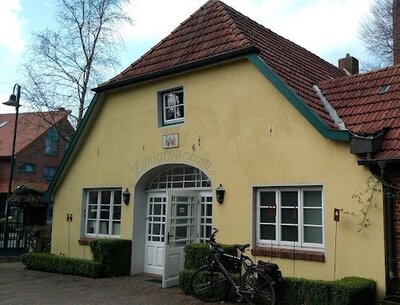 Gelbes kleines Gebäude mit rotem Ziegeldach und grün abgesetzten Kanten; mittig eine weiße Doppeltür in einem rundbogigen Eingangsbereich, zwei Fensterfronten, ein abgestelltes Fahrrad vor niedrigen Hecken sowie kahle Bäume und eine Straßenlaterne im Hintergrund.