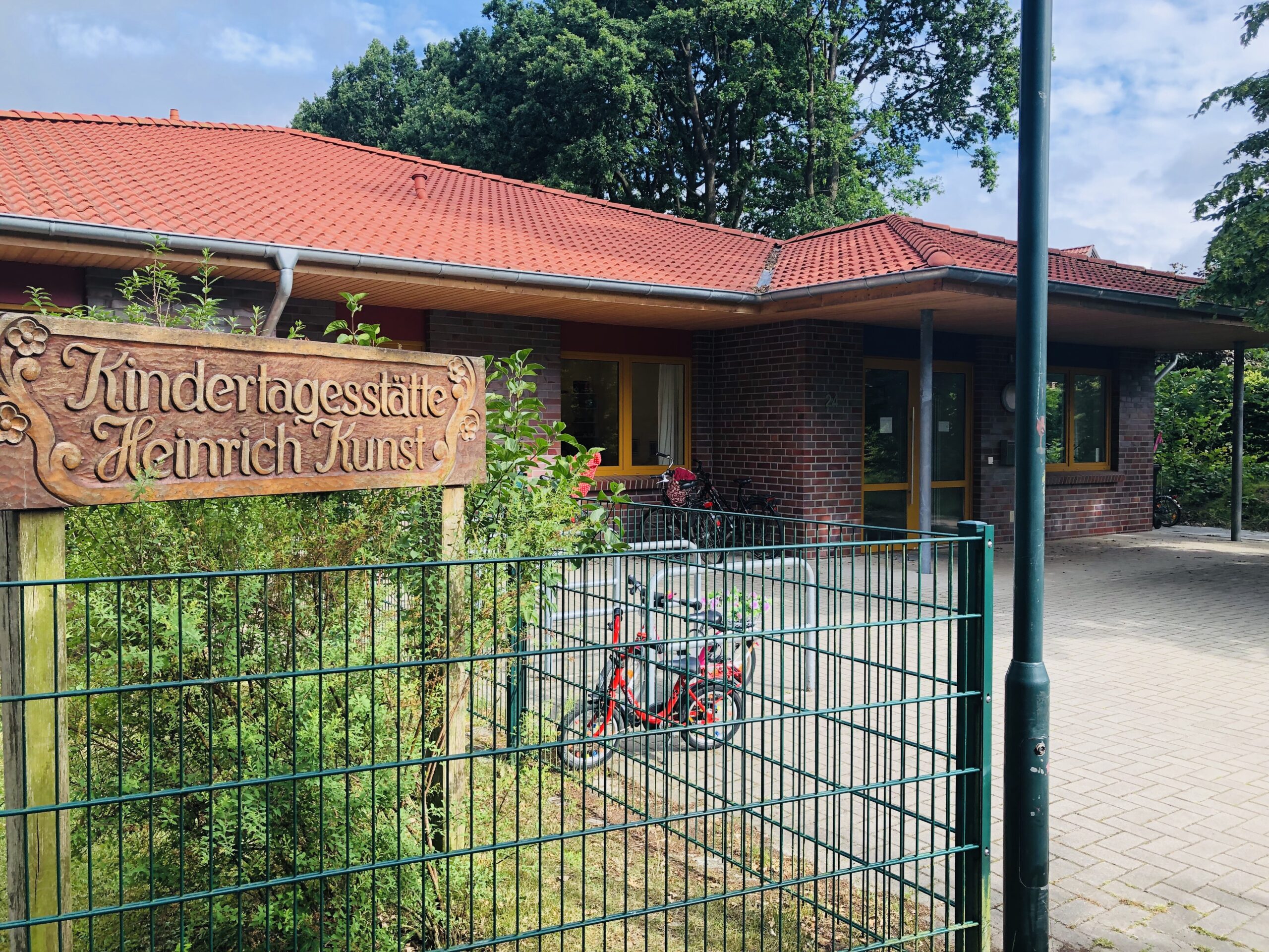 Eingangsbereich einer Kindertagesstätte mit geschnitztem Holzschild »Kindertagesstätte Heinrich Kunst« am grünen Metallzaun, dahinter Backsteingebäude mit rotem Ziegeldach, Fahrrädern und überdachtem Eingangsbereich.