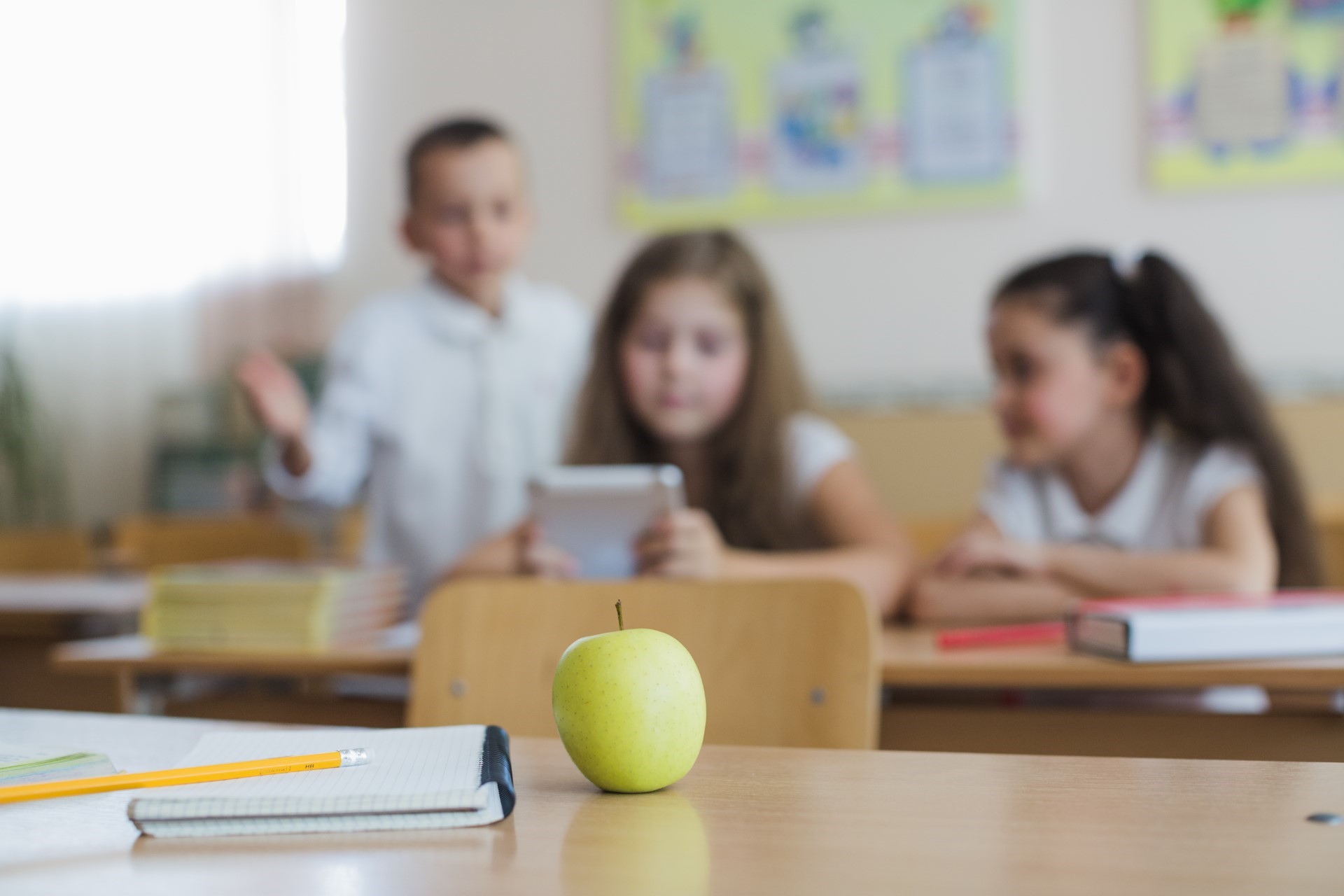 Grüner Apfel liegt auf einem Schulpult im Vordergrund; im unscharfen Hintergrund sitzen drei Kinder in einem Klassenzimmer mit Heften, Bleistift und einem Tablet.