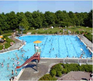 Außen-Schwimmbad mit großem rechteckigem Becken mit Schwimmbahnen, daneben ein flacheres Kinderbecken mit roter Rutsche und Lifeguard-Sitz mit gelbem Sonnenschirm; zahlreiche Badegäste im Wasser und auf Liegewiesen vor einer Baumreihe.