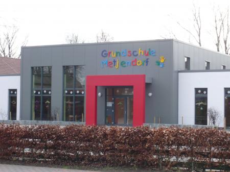 Vorderansicht einer modernen Grundschule mit dunkelgrauer Fassade, einem markanten roten Eingangsportal, buntem Schriftzug 'Grundschule' über der Tür und einer niedrigen Hecke im Vordergrund.