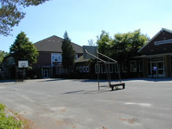 Leerer Schulhof mit großer Asphaltfläche, zwei Basketballkörben und eingeschossigem Schulgebäude im Hintergrund, umgeben von Bäumen unter klarem blauem Himmel.