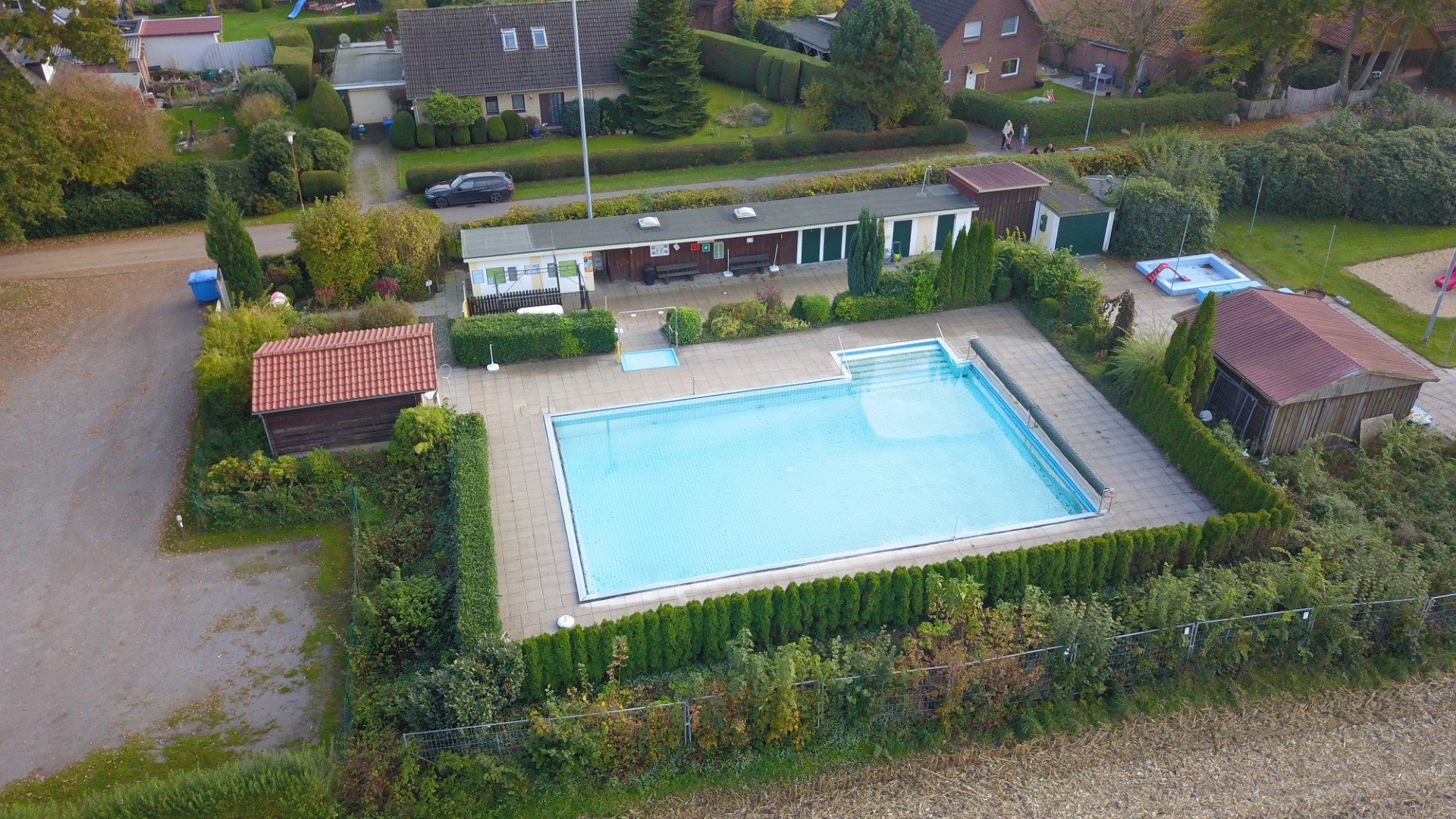 Vogelperspektive auf ein rechteckiges Freibad mit klarem blauem Wasser und Einstiegstreppen, umgeben von gepflasterter Liegefläche, dichten Hecken und mehreren Holzschuppen; im Hintergrund Wohnhäuser, Gärten und ein kleiner Spielbereich.