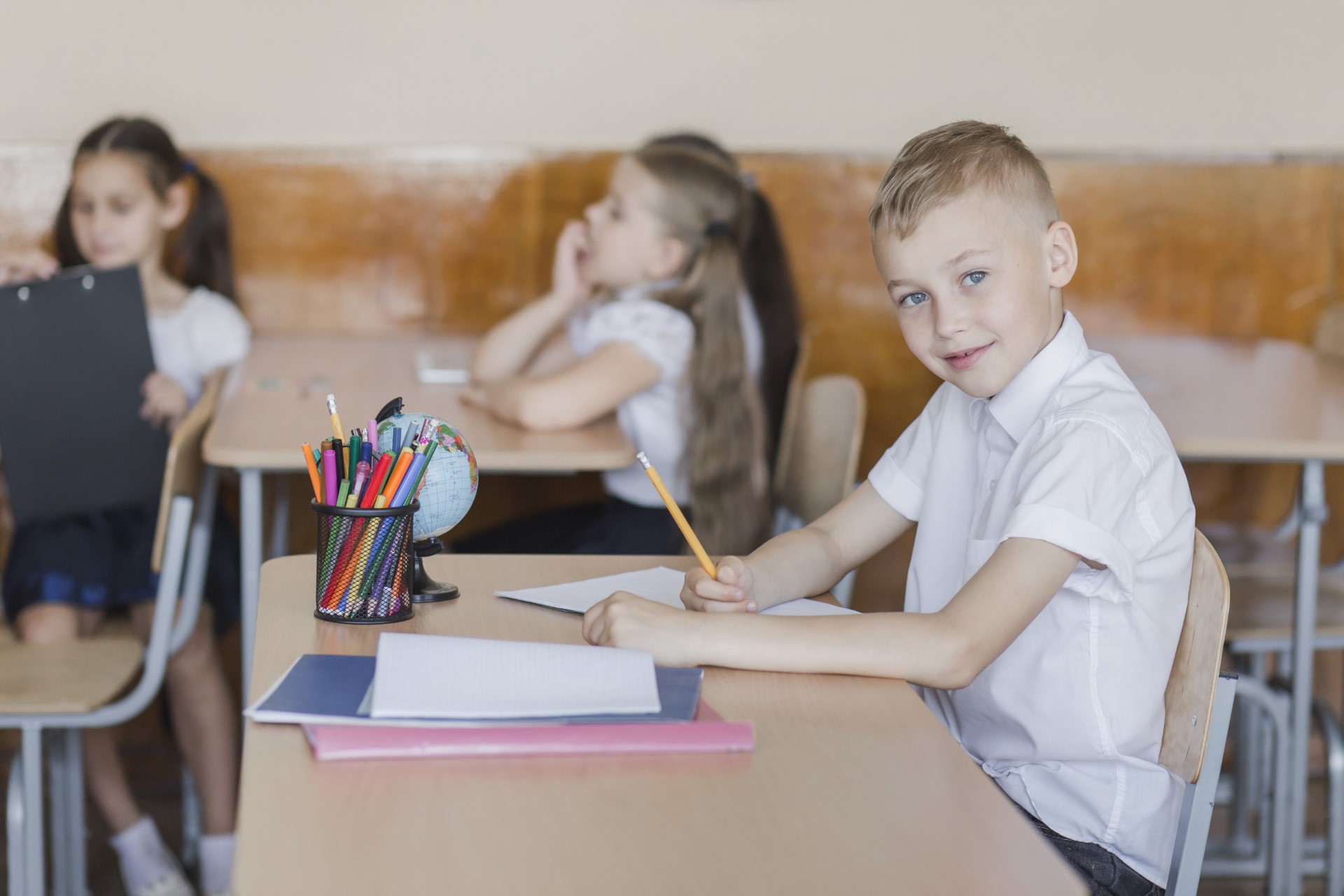 Ein lächelnder Junge mit kurzem blondem Haar in weißem Hemd sitzt an einem Klassentisch, hält einen Bleistift über ein Blatt; vor ihm liegen Hefte, ein Stiftebehälter mit bunten Stiften und ein kleiner Globus, im Hintergrund unscharfe Mitschülerinnen.