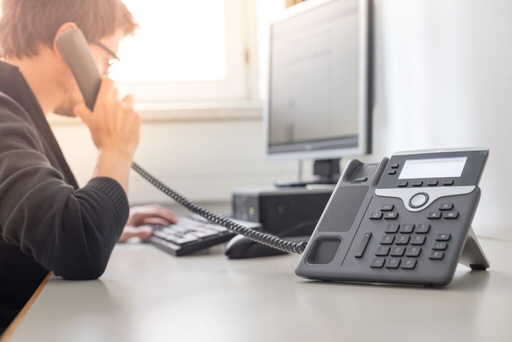 VoIP-Tischtelefon im Vordergrund; im Hintergrund eine Person am Arbeitsplatz, die den Hörer ans Ohr hält und an einer Tastatur vor einem Monitor arbeitet.