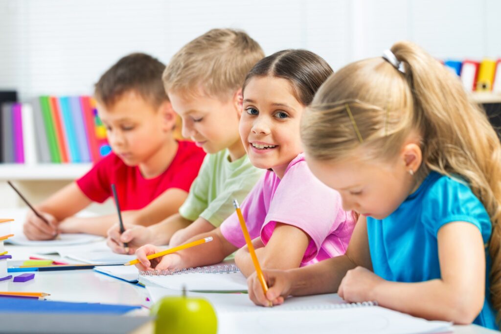 Vier Schulkinder sitzen nebeneinander an einem Tisch und schreiben mit Bleistiften in Spiralblöcke; ein Mädchen im rosa T‑Shirt lächelt in die Kamera, im Hintergrund bunte Ordner und im Vordergrund ein grüner Apfel.