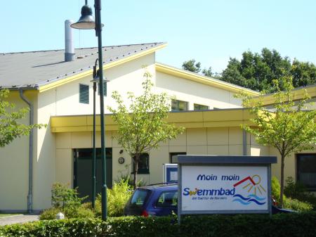 Hellgelbes, modernes Schwimmbadgebäude mit Vordach; im Vordergrund ein Schild mit der Aufschrift Moin moin Schwimmbad und stilisiertem Sonnen- und Wellenlogo, umgeben von kleinen Bäumen, einem Laternenmast und einem geparkten Auto.