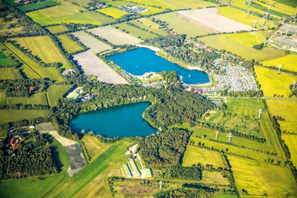 Luftaufnahme einer ländlichen Agrarlandschaft mit zwei tiefblauen Seen, dichten Bauminseln, rechteckigen Feldern, einem großen Campingplatz mit vielen Wohnwagen und verstreuten Gebäuden sowie sichtbaren Hochspannungsleitungen.