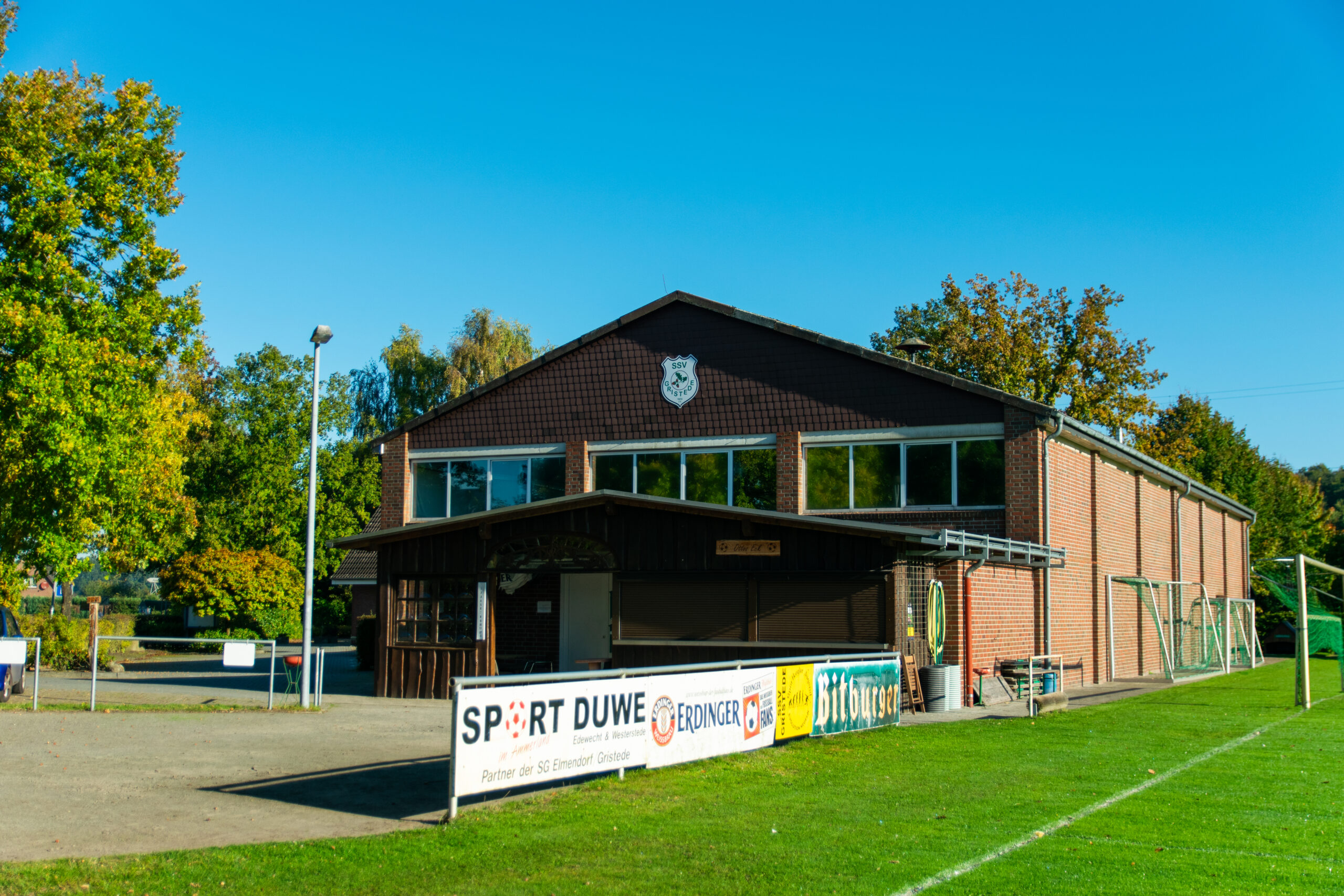 Backstein-Sportheim mit Vereinswappen, geschlossenem Schankhäuschen und Werbebanden (u. a. Sport Duwe, Bitburger) direkt an einem sonnigen Fußballrasen, im Hintergrund Bäume und blauer Himmel.