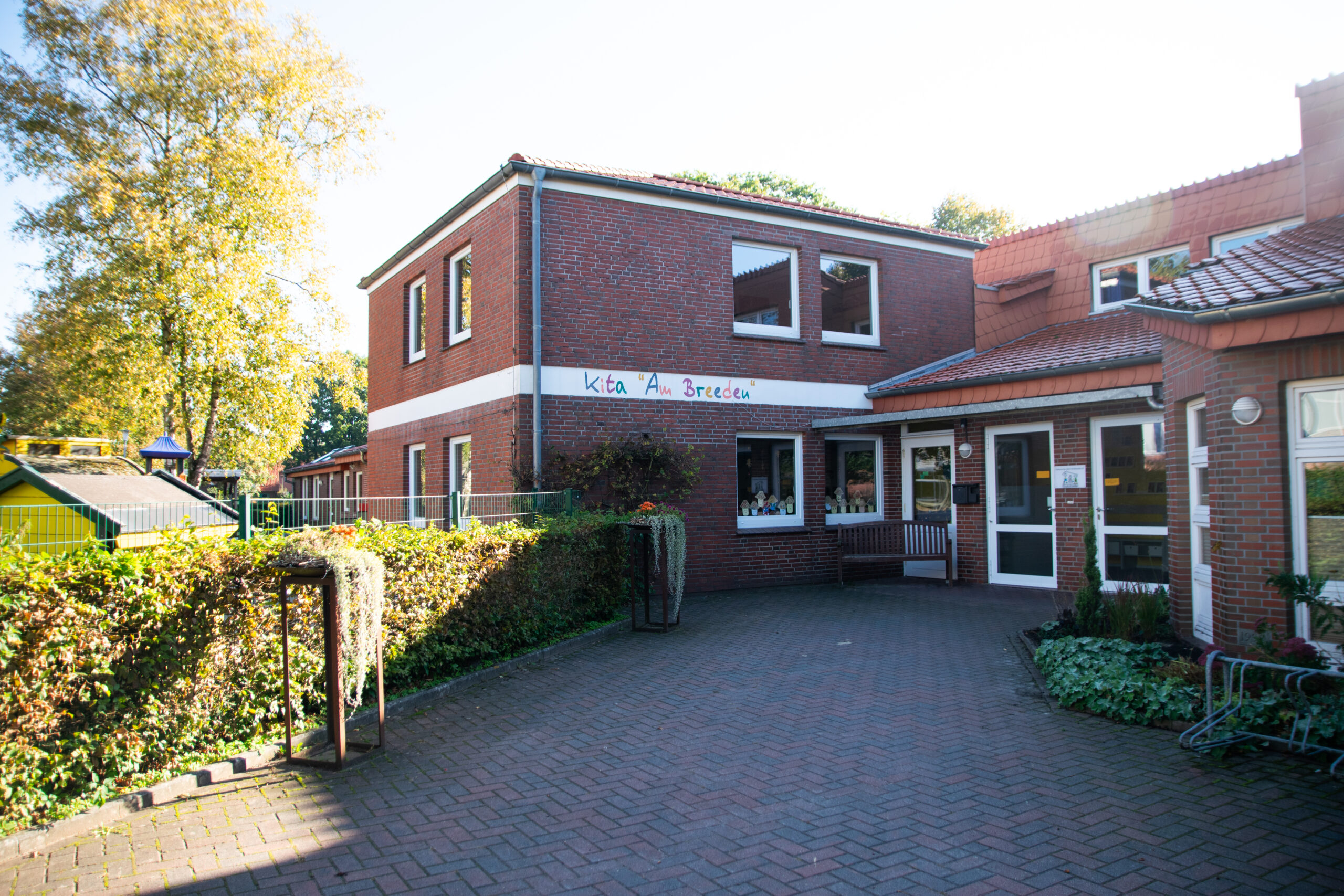 Zweistöckiges rotes Backsteingebäude einer Kindertagesstätte mit weißem Band und Schriftzug „Kita ‚Am Breeden‘“, Eingangsbereich mit Bank und Glastür, Fenster mit Kinderbasteleien, gepflastertem Vorplatz, begrüntem Zaun sowie Spielhaus und Bäumen im linken Hintergrund.