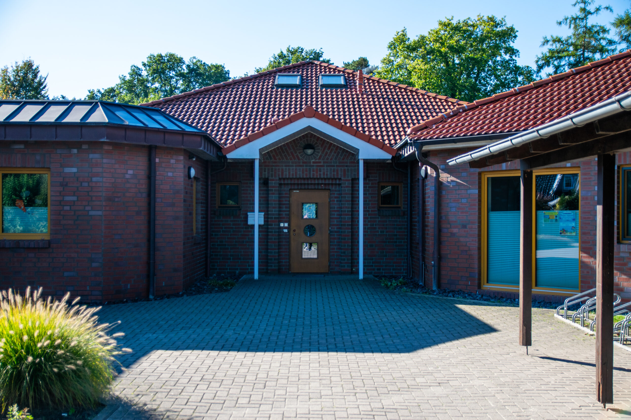 Eingangsbereich eines eingeschossigen Backsteingebäudes mit rot gedecktem Dach und zwei Dachfenstern, zentraler Holztür mit drei kleinen Fenstern unter weißem Giebel, gepflastertem Vorplatz, Ziergras links und Fahrradständern rechts.
