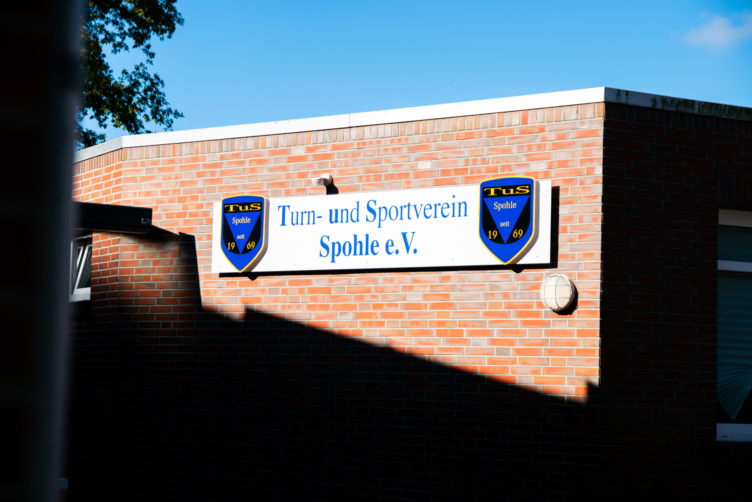 Rote Backsteinfassade mit einem weißen Schild des Turn- und Sportvereins Spohle e.V., flankiert von zwei blauen Vereinswappen und teils von einem schrägen Schatten sowie einer Außenlampe bedeckt.