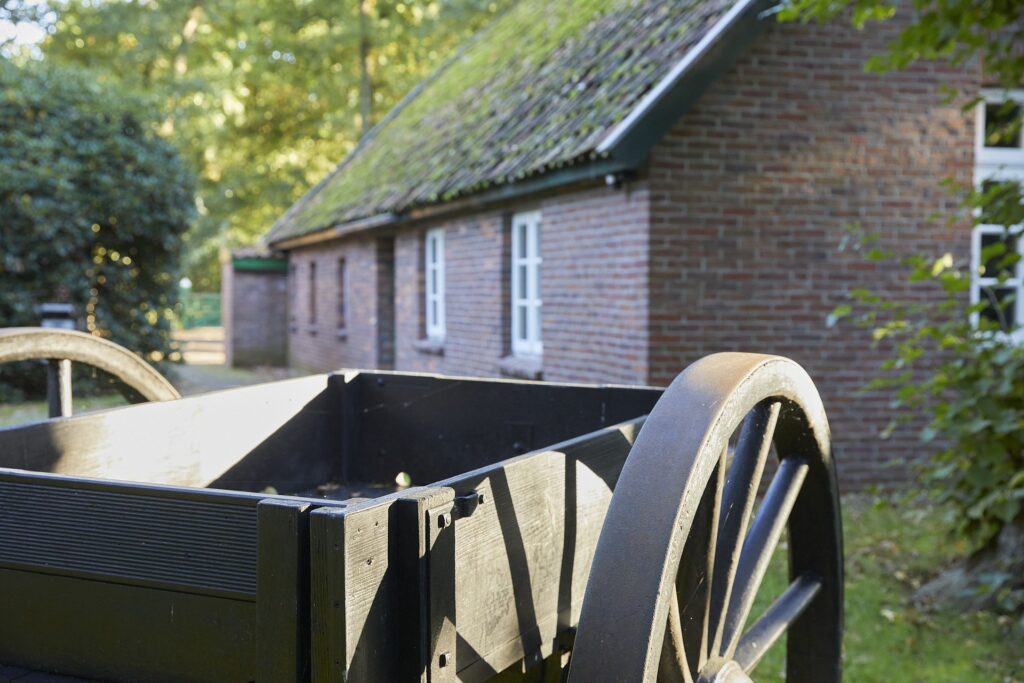 Ein schwarzer hölzerner Handwagen mit großem Metallrad im Vordergrund, dahinter ein Ziegelsteinhaus mit weißen Fenstern, moosbedecktem Dach und von Sonnenlicht durchfluteten Bäumen.