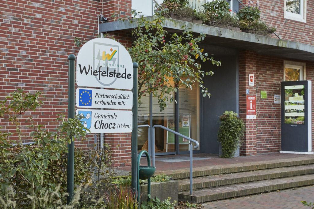 Ein Ortsschild für Wiefelstede steht vor einem Backsteingebäude mit Treppenaufgang und Pflanzen; darunter Zusatztafeln mit EU-Flagge und dem Hinweis auf die Partnerschaft zur Gemeinde Chocz (Polen).