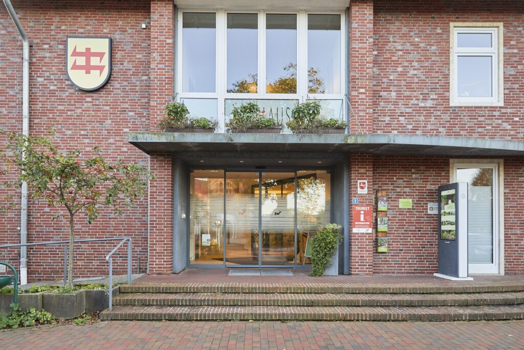 Eingangsbereich eines roten Backsteingebäudes mit mehrstufiger Treppe, Handlauf und kleinem Baum links, automatischen Glastüren, darüber Fenstern mit Blumenkästen, einem Wappenschild an der Fassade links sowie einem Informationsautomaten und Tourist‑Info‑Schild rechts.