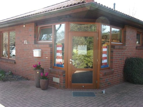 Eingang eines eingeschossigen Backsteinhauses mit brauner Holzrahmen-Glas-Eingangstür, Hausnummer 17a, weißem Briefkasten links, zwei Topfpflanzen mit pinken Blüten, rot-weißen Streifen an den Seitenscheiben und gepflastertem Vorplatz.