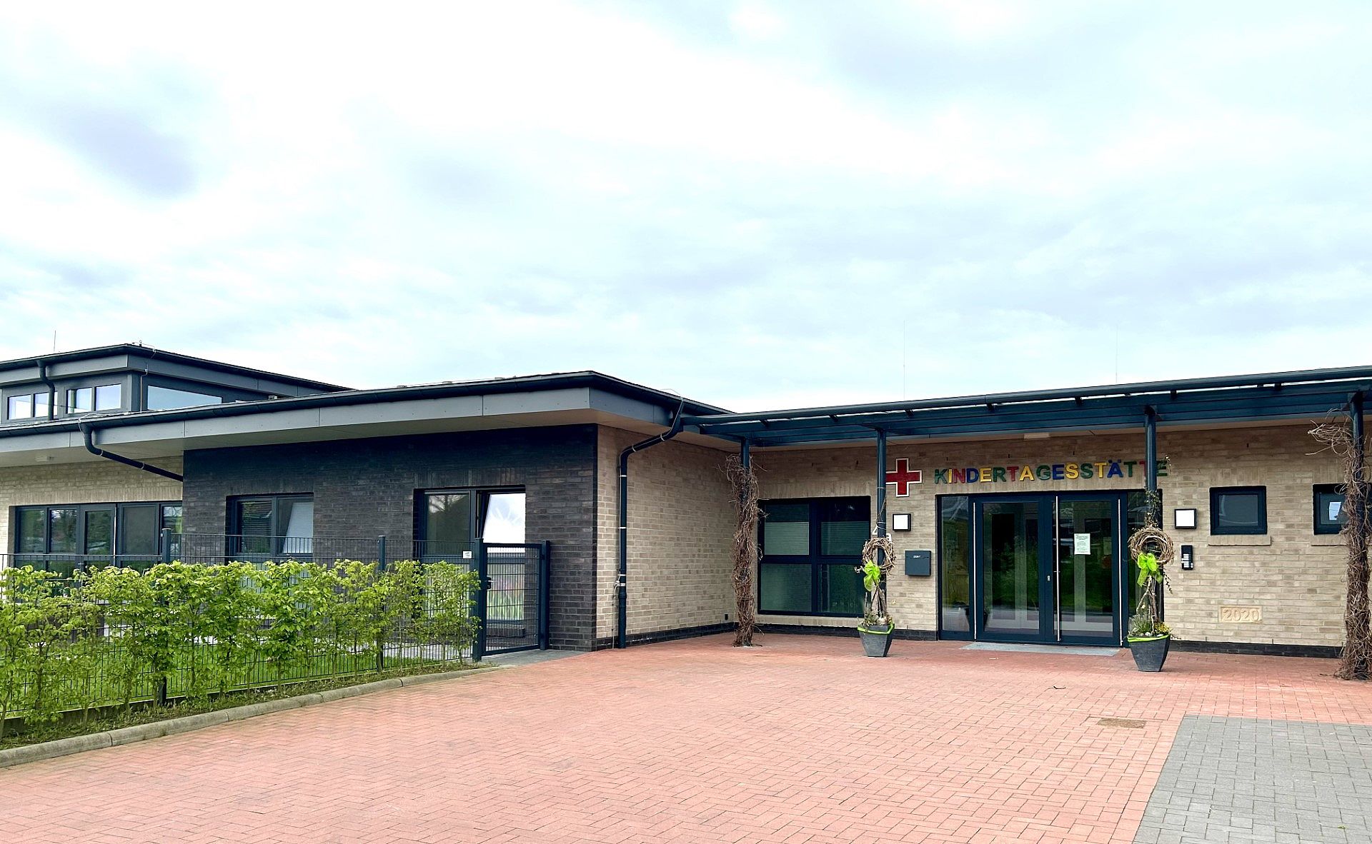 Eingangsbereich einer modernen Kindertagesstätte mit beigefarbener Backsteinfassade, farbigem Schild "KINDERTAGESSTÄTTE" neben einem roten Kreuz, doppelten Glastüren, zwei geschmückten Pflanzkübeln und gepflastertem Vorplatz, links eine niedrige Hecke und Zaun.