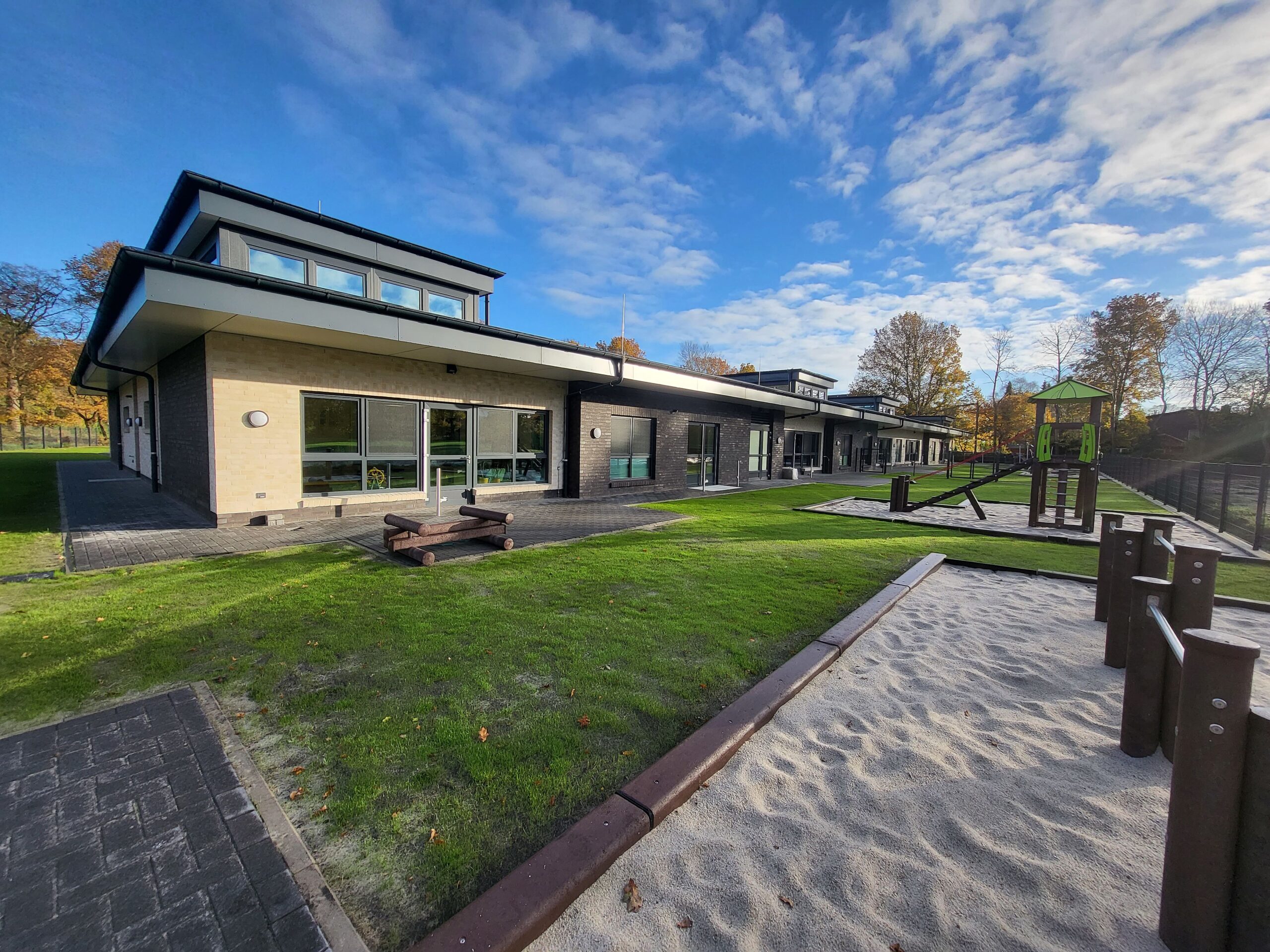 Moderner eingeschossiger Gebäudekomplex mit hellen und dunklen Fassaden, großen Fensterfronten und flachem Vordach, davor gepflegte Rasenflächen, Sandflächen und ein hölzernes Kletter- und Spielgerät mit grünem Dach unter blauem Herbsthimmel.