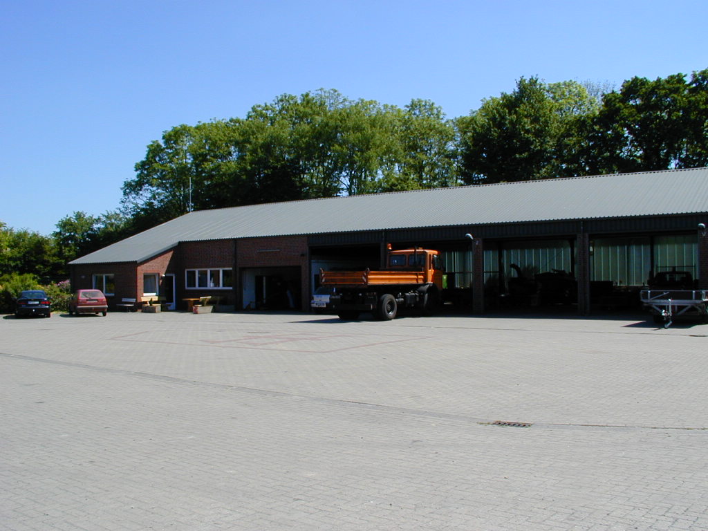 Langer niedriger Backsteinbetrieb mit Wellblechdach an einem hellen, gepflasterten Hof; vor den offenen Toren steht ein orangefarbener Lkw, links zwei Pkw, im Hintergrund hohe Bäume unter klarem blauem Himmel.