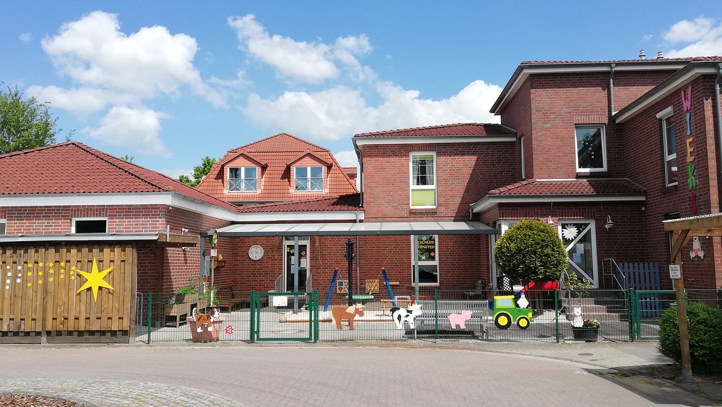Rotes Backsteingebäude einer Kindertagesstätte mit überdachtem Eingang, eingezäuntem Spielhof mit bunten Holz-Tierfiguren, einer großen Stern-Dekoration an einem Holzzaun und blauem Himmel mit weißen Wolken.