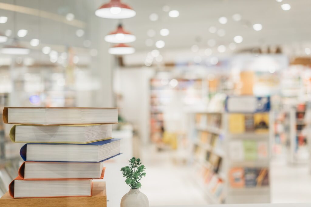 Links im Vordergrund ein Stapel Bücher mit bunten Einbänden auf einem Tisch und eine kleine weiße Vase mit grünem Pflänzchen, im Hintergrund unscharfe Regale und hängende Lampen einer hellen modernen Buchhandlung.