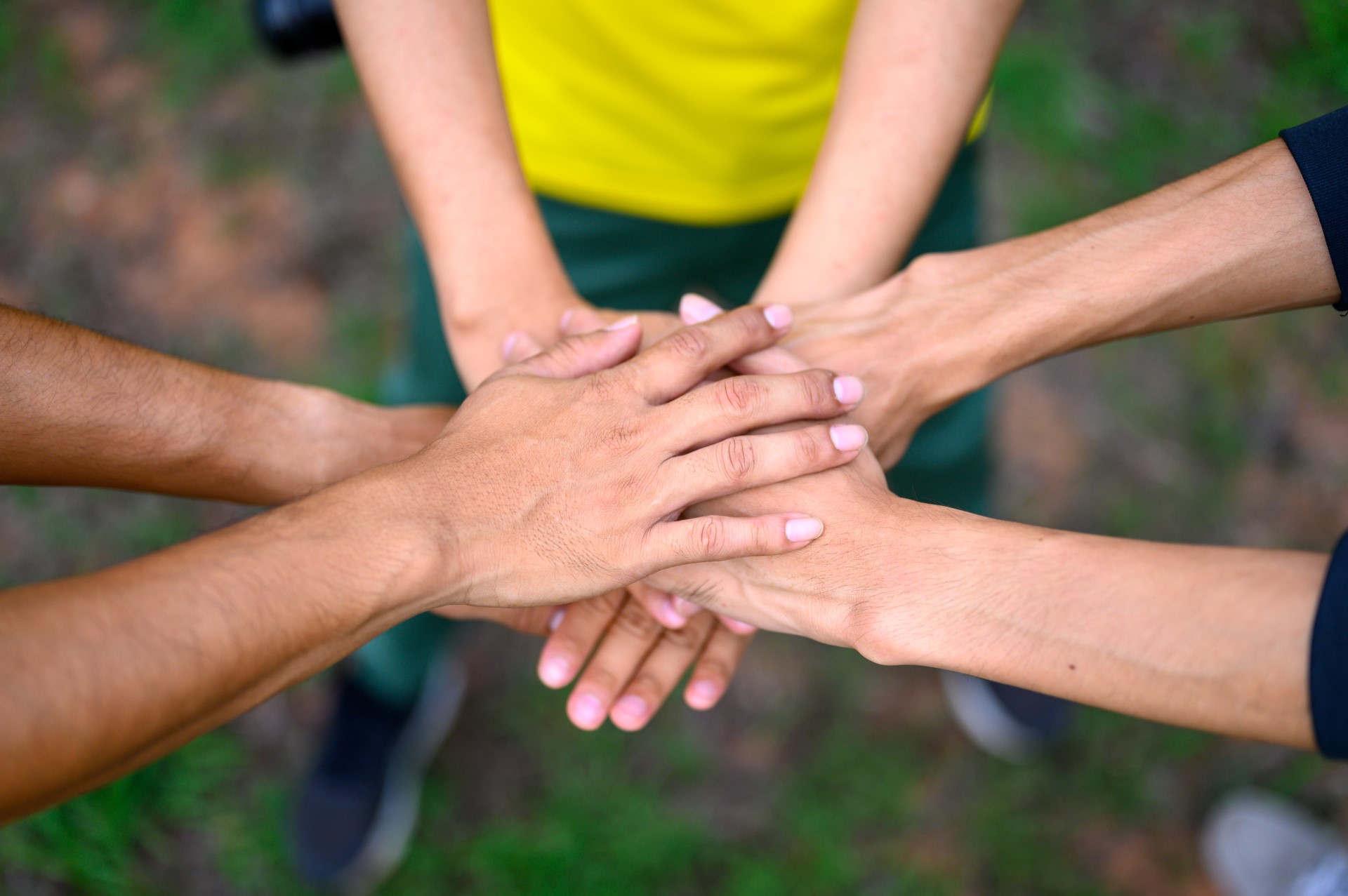 Mehrere Hände unterschiedlicher Personen übereinandergelegt in einer Teamgeste, mit sichtbarem gelben Shirt und unscharfem grünem Außenhintergrund.