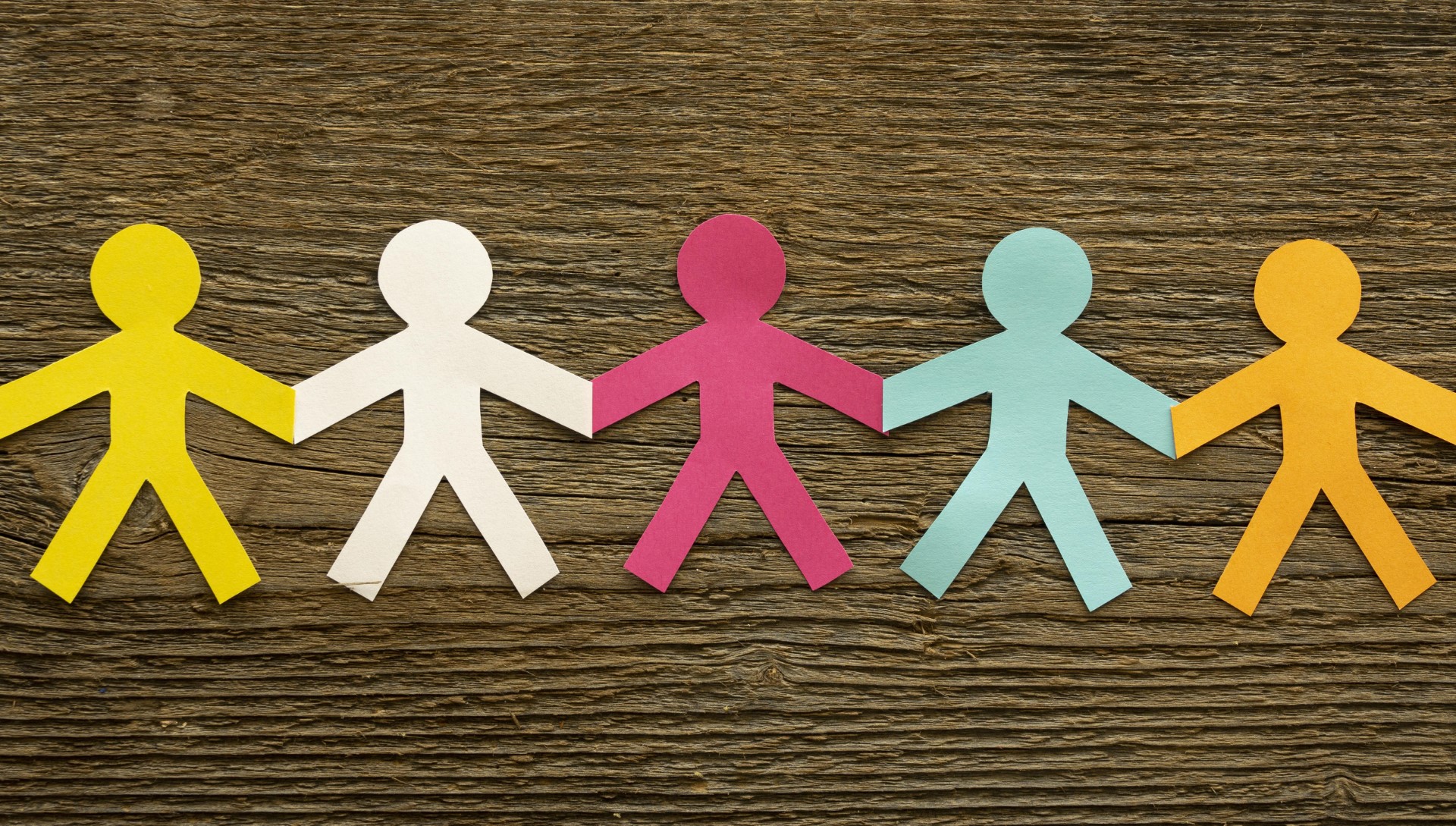 Fünf farbige Papierfiguren (gelb, weiß, rosa, hellblau, orange) als Menschenkette an den Händen verbunden auf einer groben Holzoberfläche.