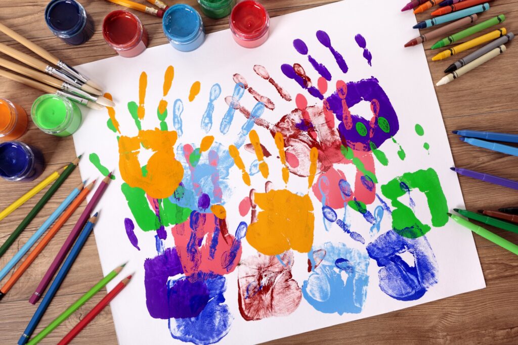 Weißes Blatt mit zahlreichen überlappenden, bunten Handabdrücken in Orange, Blau, Grün, Violett und Rot, auf einem Holztisch platziert und umgeben von geöffneten Farbtöpfchen, Pinseln, Buntstiften und Wachsmalkreiden.