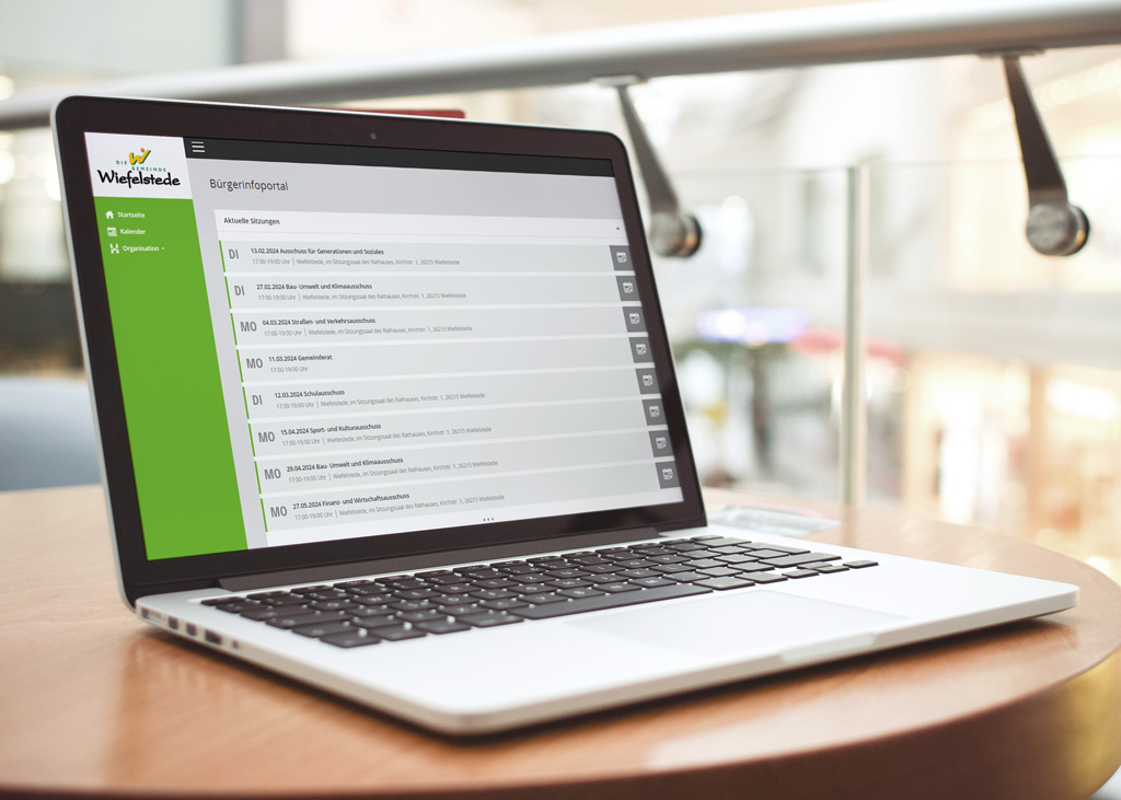 Auf einem runden Holztisch in einem hellen Innenraum steht ein aufgeklappter Laptop, dessen Bildschirm das Bürgerportal der Gemeinde Wiefelstede mit grünem Seitenmenü und einer Liste aktueller Sitzungen anzeigt.