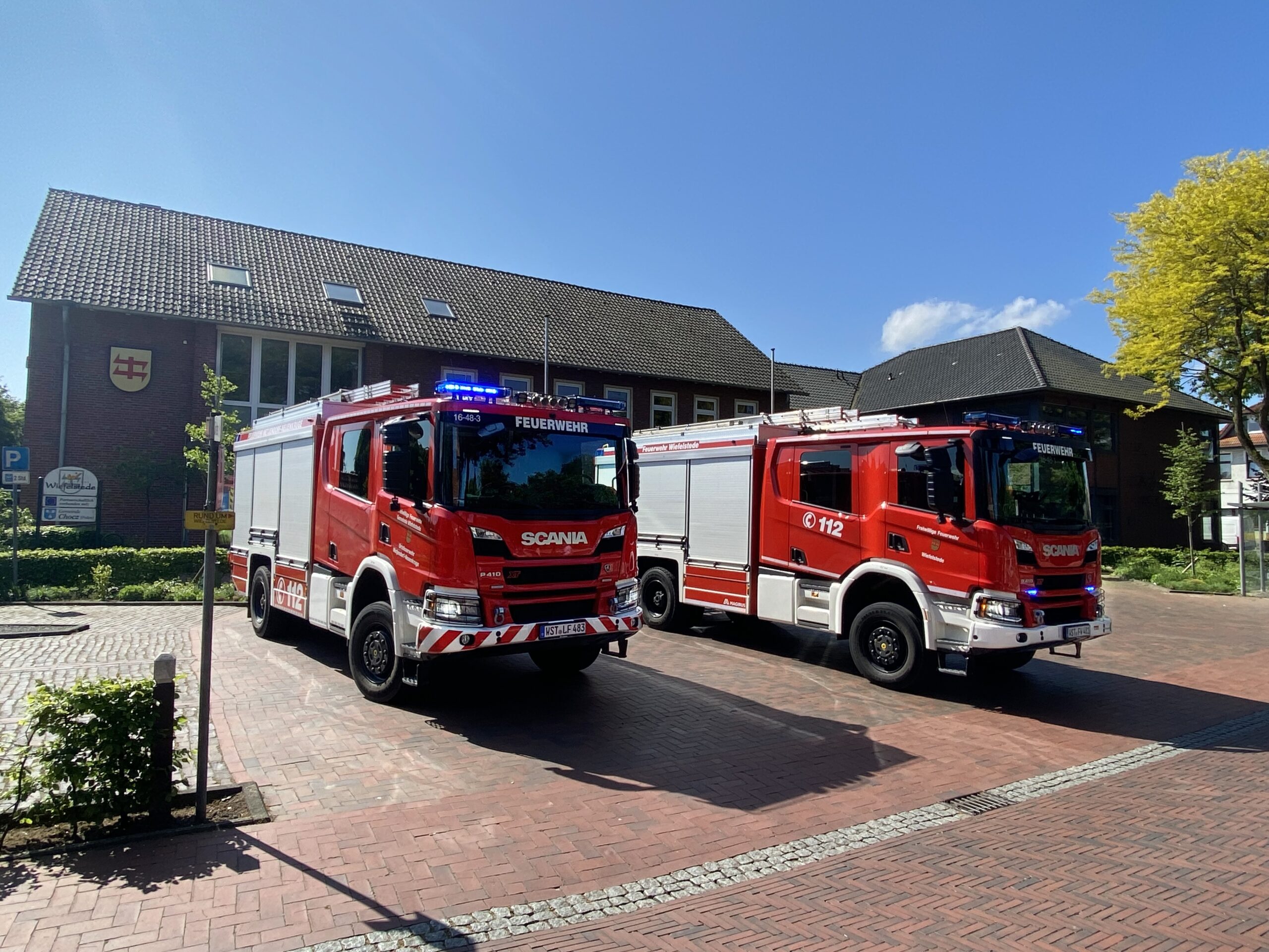 Zwei nebeneinander geparkte rote Feuerwehrfahrzeuge der Marke Scania mit eingeschalteten blauen Warnleuchten und seitlicher 112‑Kennzeichnung auf einem gepflasterten Vorplatz vor einem Backsteingebäude mit Wappenschild unter klarem blauem Himmel.