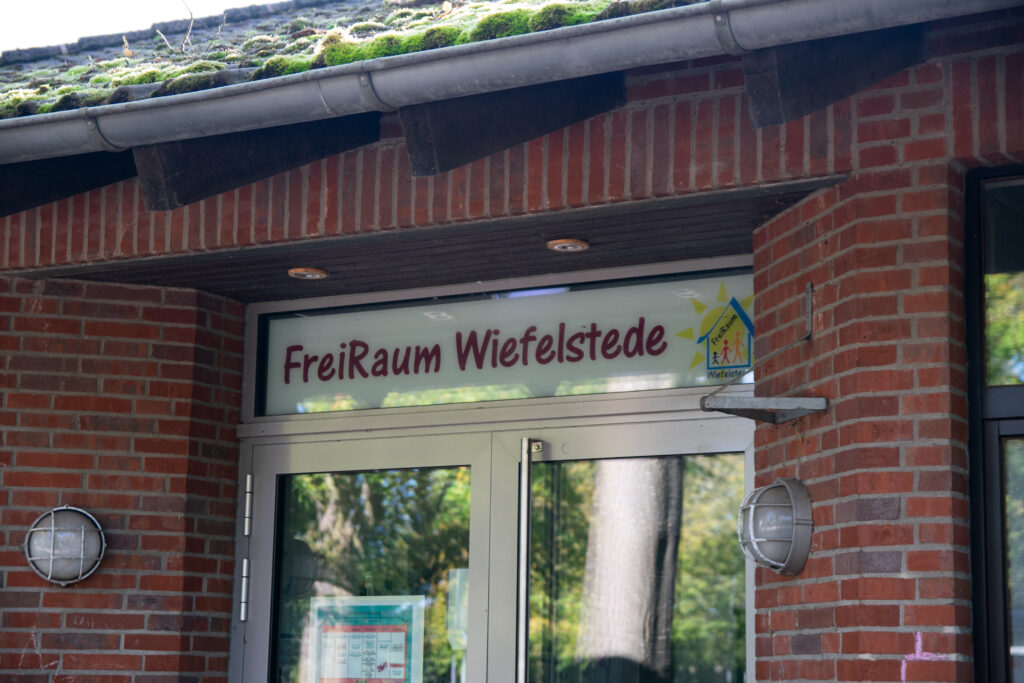 Eingang eines roten Backsteingebäudes mit Glastür, über der Tür das Schild ‚FreiRaum Wiefelstede‘, seitlichen Wandlampen und einem moosbedeckten Dach.