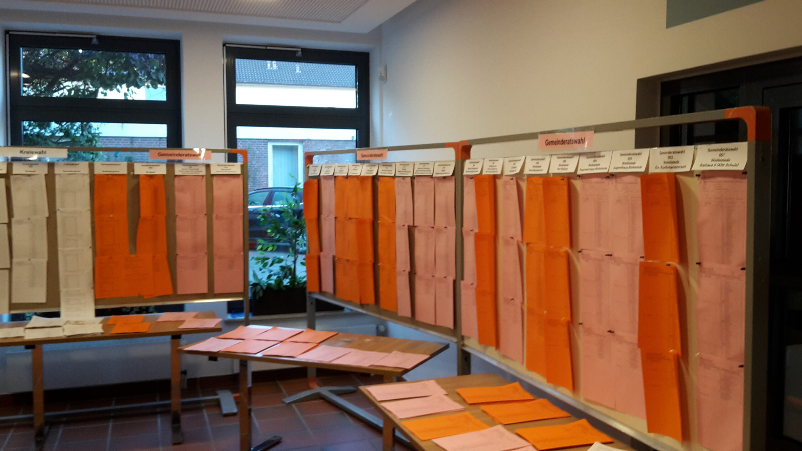 Stellwände in einem Wahlraum, an denen Reihen orange und rosa Wahlzettel und Listen befestigt sind, sowie Tische mit weiteren ausliegenden Blättern vor großen Fenstern.