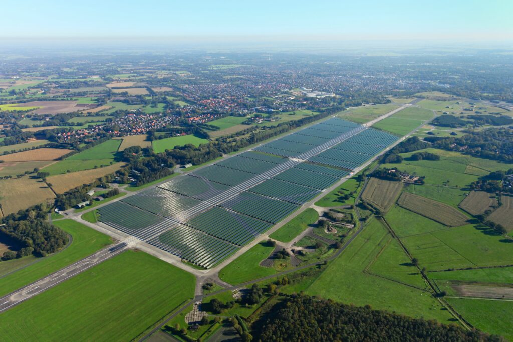 Luftaufnahme eines großen Solarparks mit in Reihen angeordneten Photovoltaikmodulen auf einem ehemaligen Flugfeld, umgeben von grünen Feldern, Wegen und einem Dorf am Horizont.