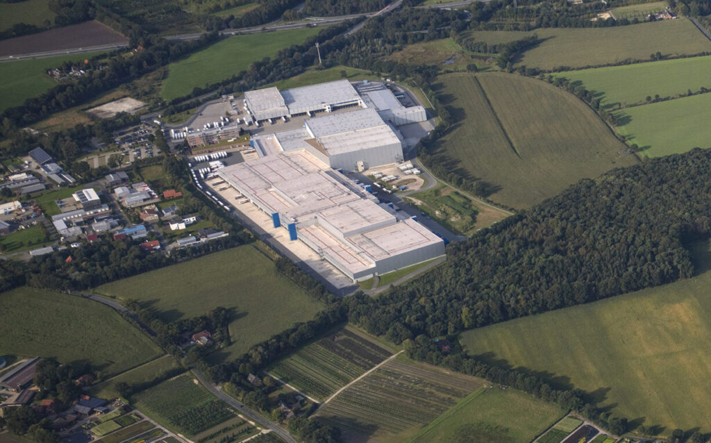 Luftaufnahme eines großen Industrie- und Logistikkomplexes mit mehreren hellen, rechteckigen Hallendächern, Laderampen und abgestellten Lkw, umgeben von landwirtschaftlichen Feldern, Waldflächen und einem kleinen Wohn- und Gewerbegebiet.