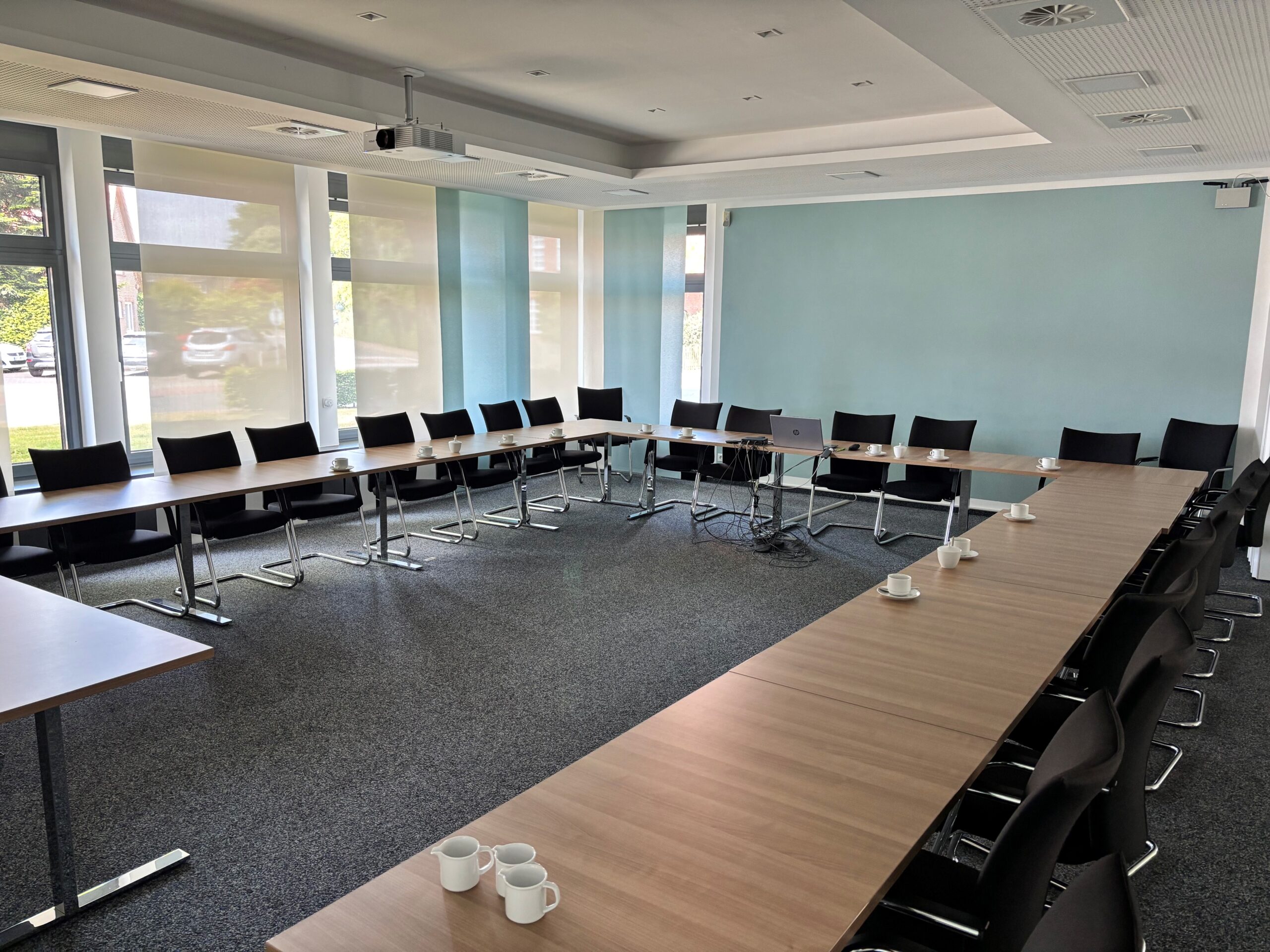 Heller Konferenzraum mit in U‑Form angeordneten Holztischen und schwarzen Stühlen, mehreren weißen Kaffeetassen, einem Laptop mit Kabeln in der Mitte, großen Fenstern mit Rollos, einer hellblauen Wand und Deckenprojektor.