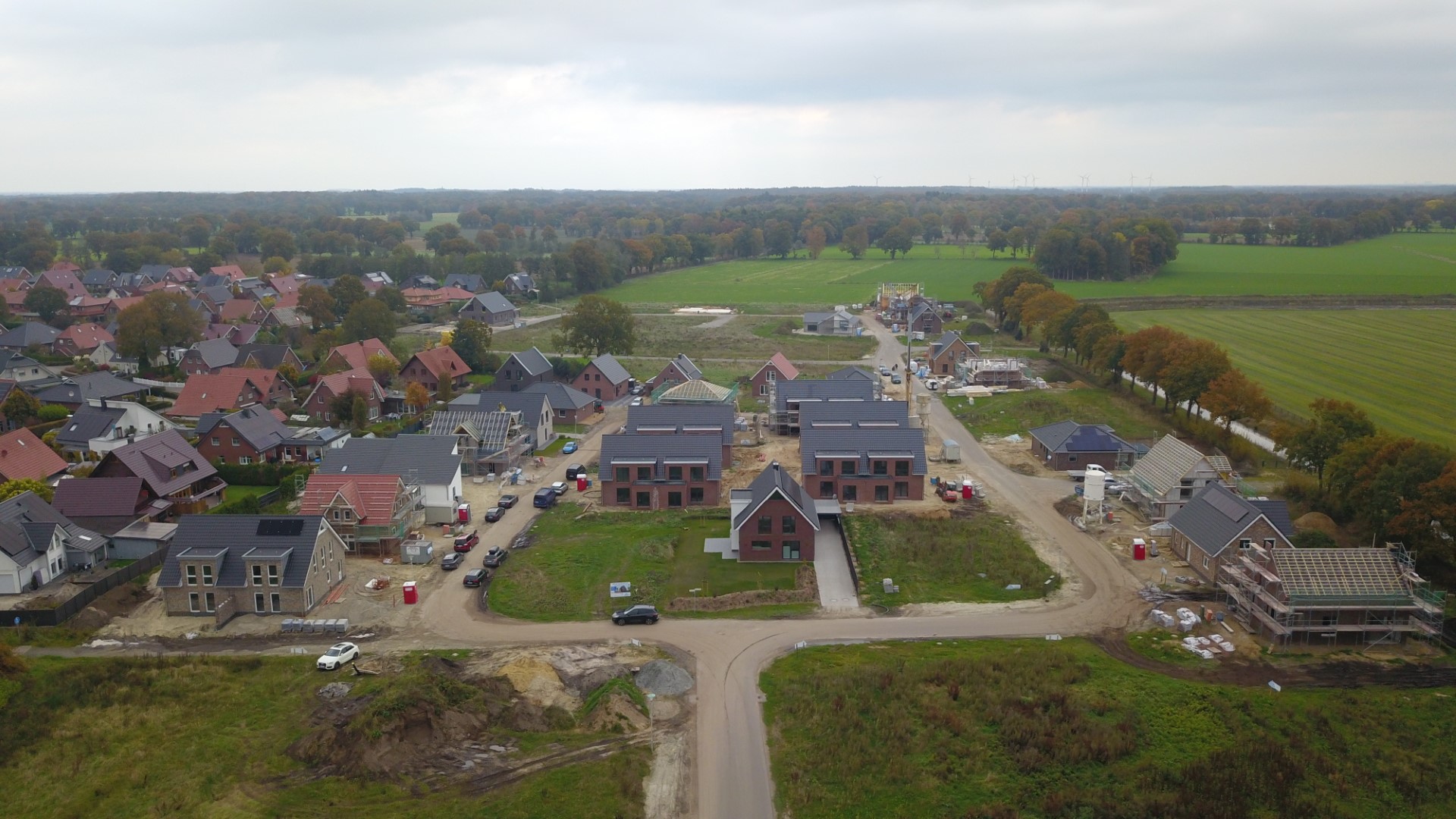 Luftaufnahme einer Neubausiedlung mit roten und grauen Einfamilienhäusern, mehreren im Bau befindlichen Gebäuden, Baustellenfahrzeugen und Schotterstraßen, umgeben von grünen Feldern und Baumreihen unter einem bewölkten Himmel.