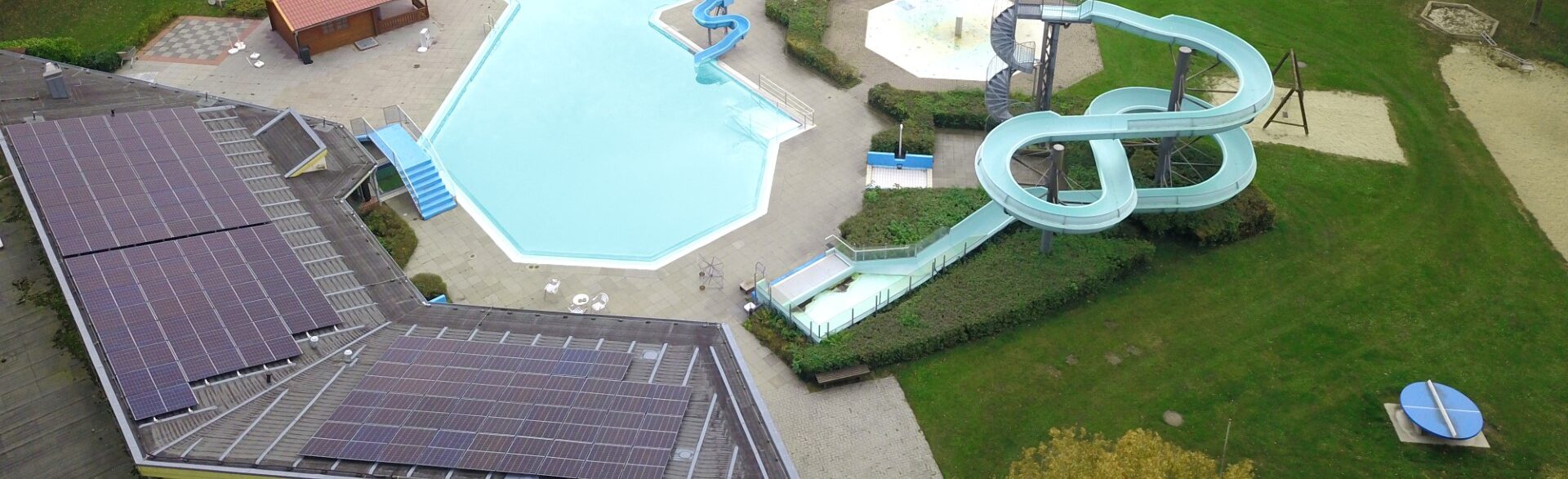 Drohnenaufnahme eines Freibads mit leerem hellblauem Schwimmbecken, geschwungenen hellgrünen Wasserrutschen, einem Gebäude mit Solarpaneelen auf dem Dach sowie umliegenden Wegen, Rasenflächen und Spielbereichen.