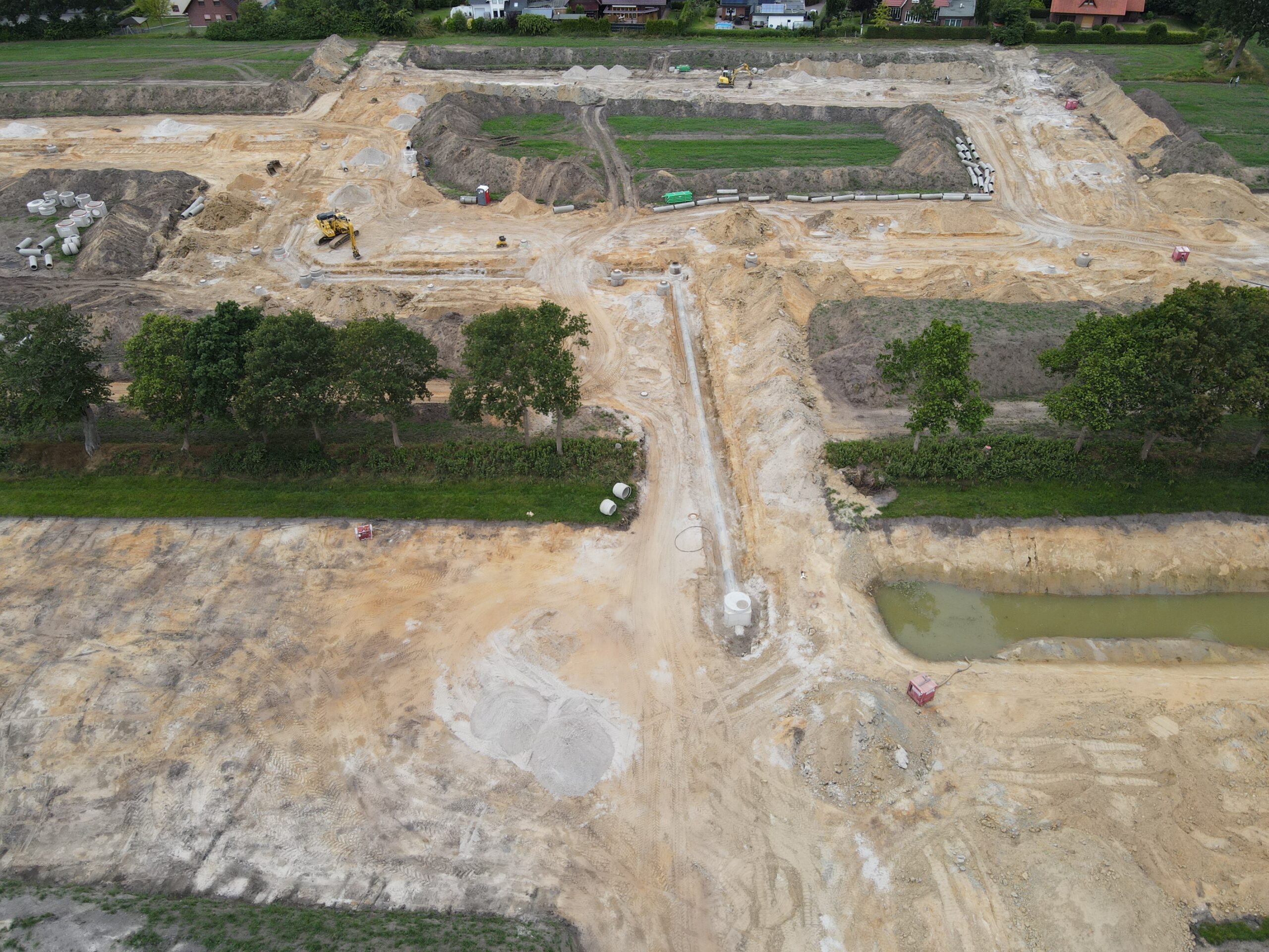 Luftaufnahme einer großflächigen Baustelle mit ausgehobenen Gräben und Sandflächen, einem Bagger, gestapelten Betonrohren, einem länglichen Wassergraben, einer Baumreihe in der Bildmitte und Wohnhäusern am oberen Bildrand.