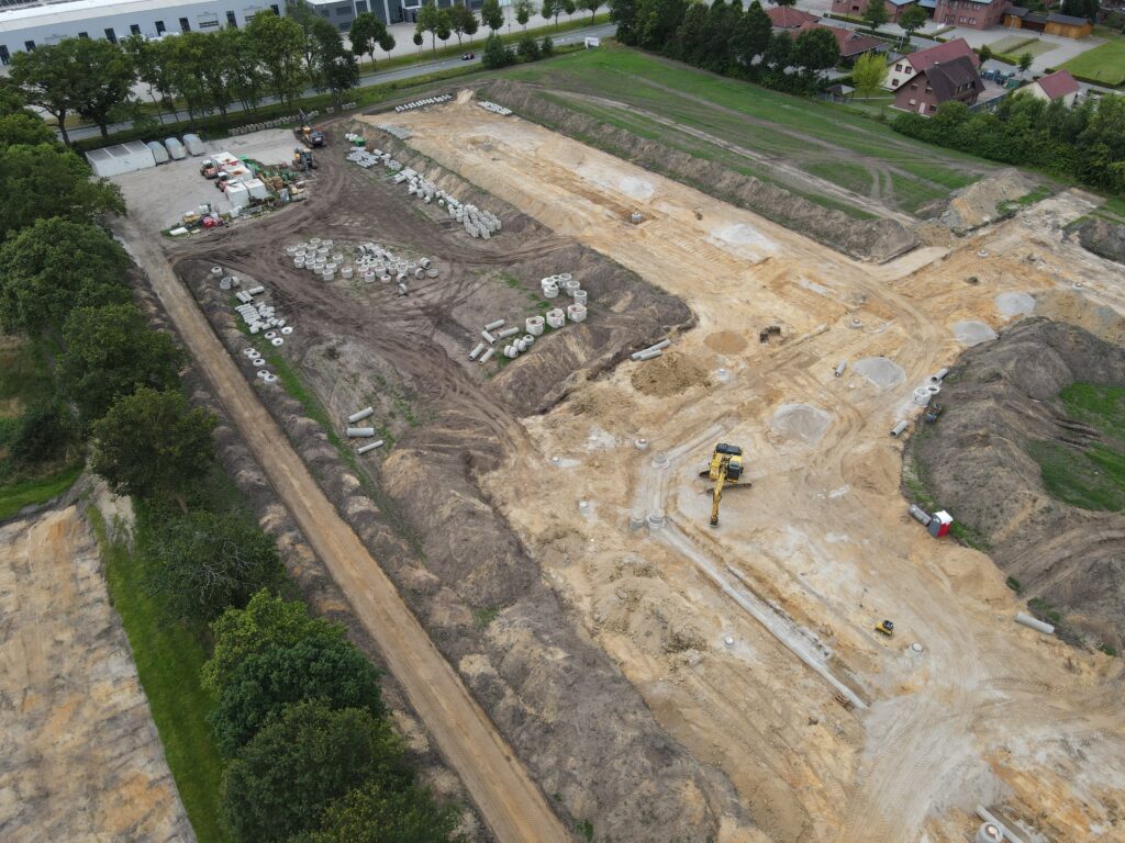 Luftaufnahme einer großen Baustelle mit ausgehobenem sandigem Boden, einem gelben Bagger in der Mitte, mehreren gestapelten Betonrohren und Baustellengeräten sowie angrenzender Baumreihe und Wohnhäusern.
