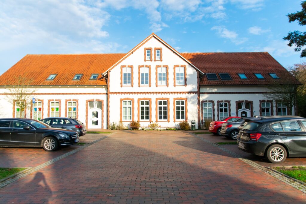 Zweistöckiges weißes Gebäude mit zentralem Giebel, rotgedecktem Ziegeldach und rot verklinkerten Fensterlaibungen, davor ein gepflasterter Hof mit mehreren geparkten Autos und blauem Himmel.