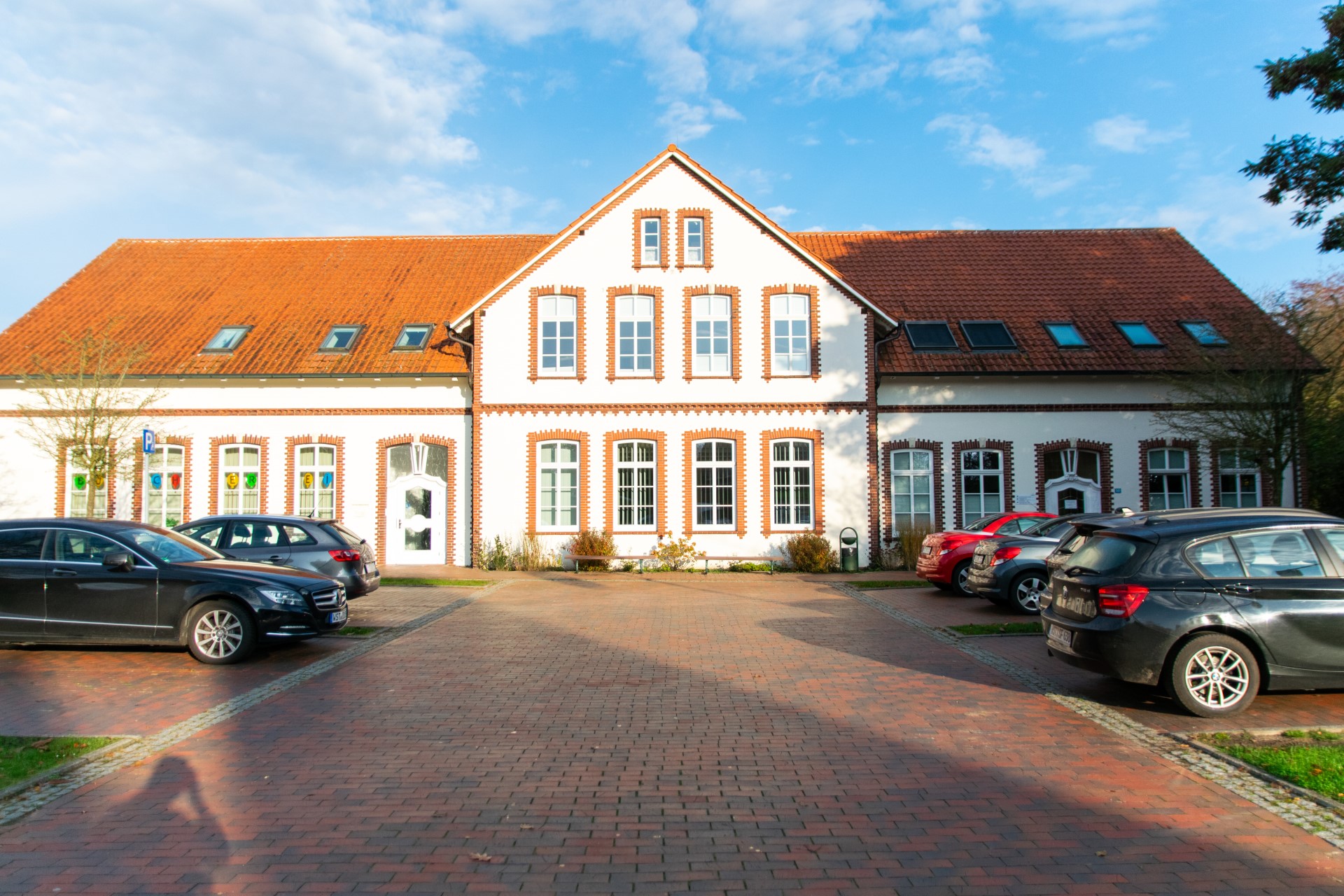 Zweistöckiges weißes Gebäude mit zentralem Giebel, rotgedecktem Ziegeldach und rot verklinkerten Fensterlaibungen, davor ein gepflasterter Hof mit mehreren geparkten Autos und blauem Himmel.