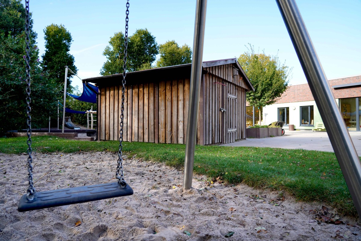 Schaukel mit schwarzem Sitz und Ketten über Sandboden im Vordergrund, dahinter ein Metallgestell, ein hölzerner Schuppen, Sonnensegel über Spielgeräten sowie ein niedriger Gebäuderiegel und Bäume im Hintergrund.