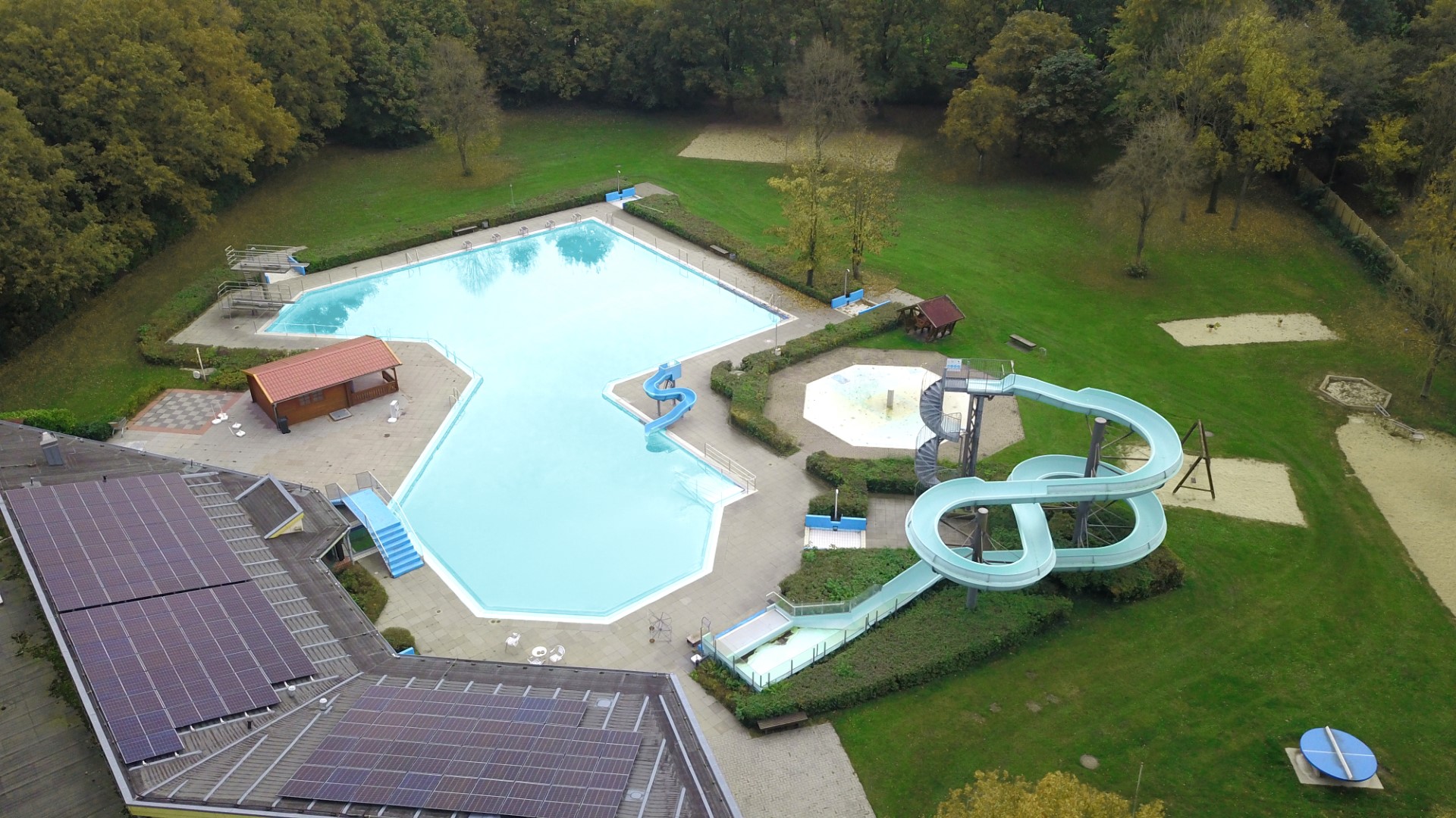 Luftaufnahme eines leerstehenden Freibadkomplexes mit großem polygonalem Außenbecken, mehreren blauen Wasserrutschen (darunter eine geschwungene Spirale), einem kleinen Holzgebäude, Solarpaneelen auf dem Hallendach sowie umgebenden Rasenflächen und Bäumen.