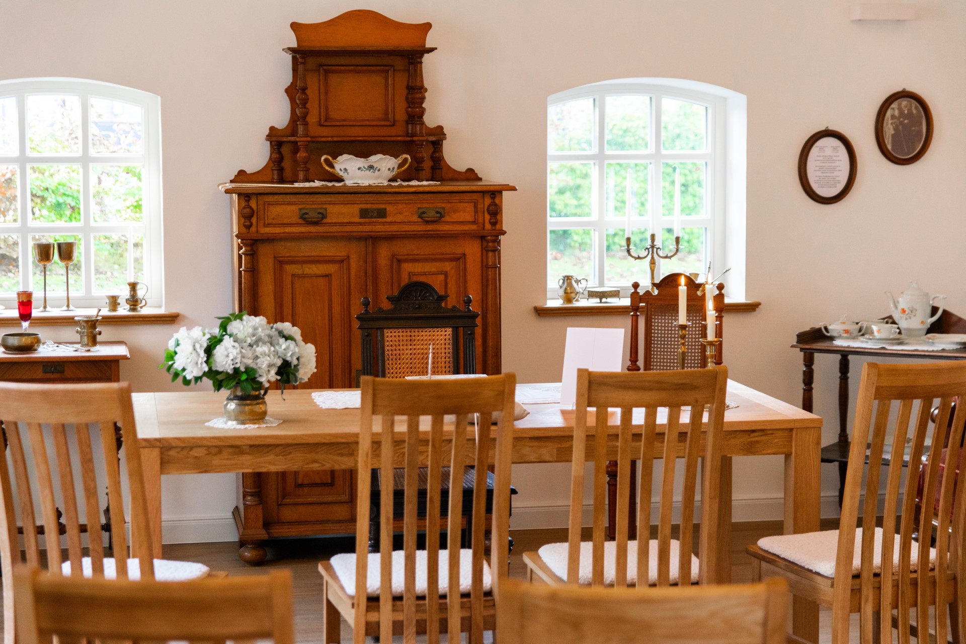 Traditioneller Essraum mit langem Holztisch und mehreren Holzstühlen, einer Vase mit weißen Blumen, brennenden Kerzen und einem großen antiken Holzbuffet vor zwei Sprossenfenstern.