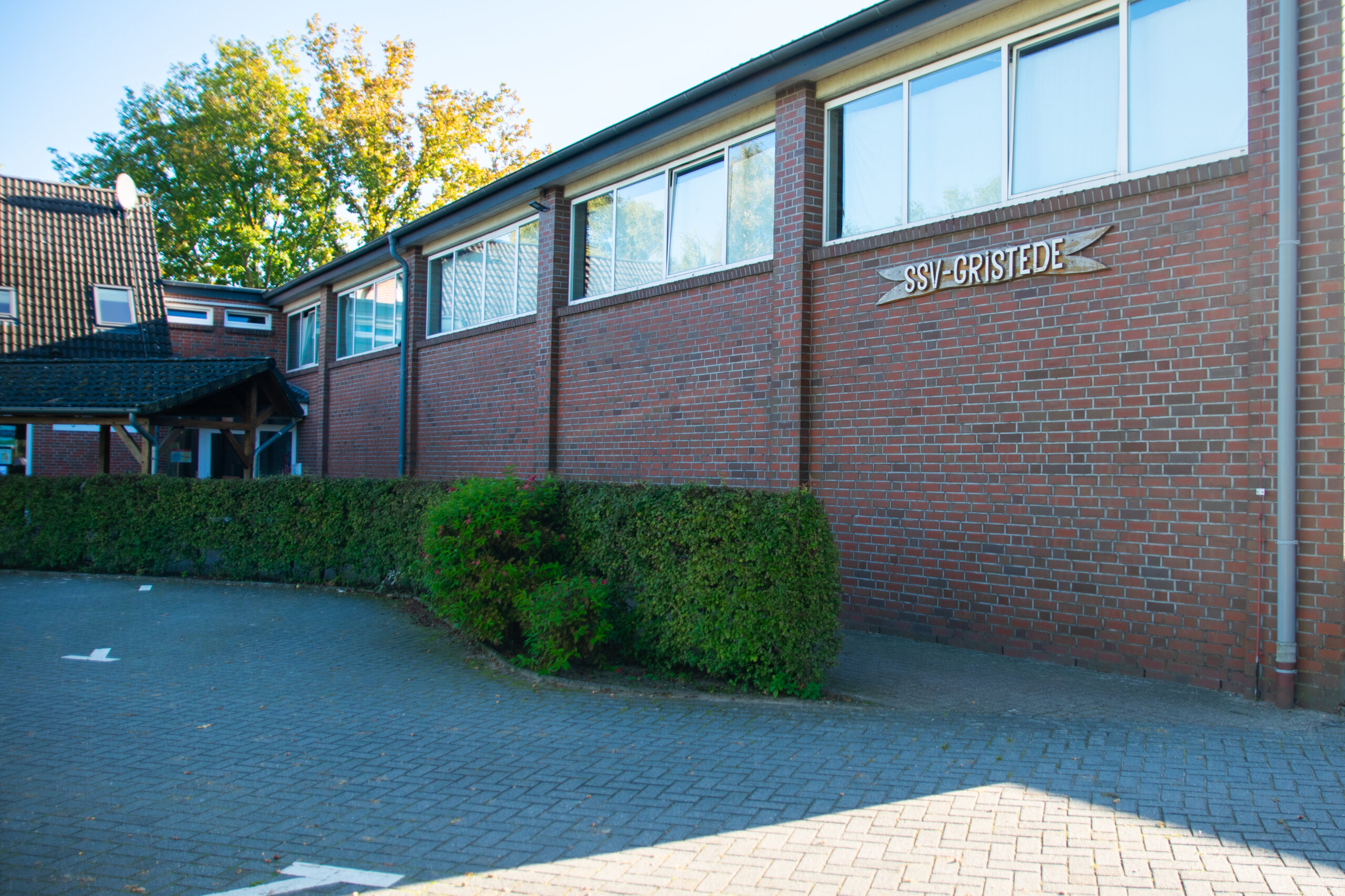 Rotes Backsteingebäude mit großen Fenstern und dem Holzschild „SSV-GRISTEDE“ an der Fassade, davor eine niedrige Hecke und ein gepflasterter Parkplatz.