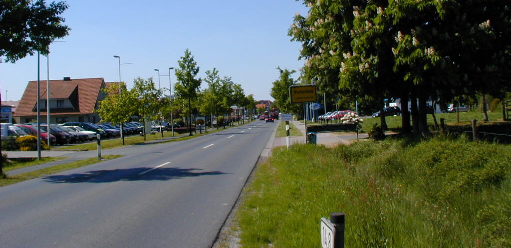Zweispurige Vorortstraße bei Sonnenschein, links Reihenparkplätze vor Häusern, rechts grasbewachsener Rand mit Bäumen und einem gelben Ortsschild, Blick in die Ferne.